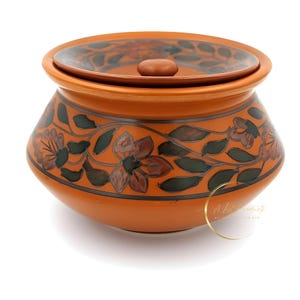Puede incluir: Un tarro de cerámica de color terracota con tapa, con un diseño floral en verde oscuro y marrón. El tarro tiene forma redondeada con tapa y un pequeño pomo. El patrón floral envuelve el cuerpo del tarro.