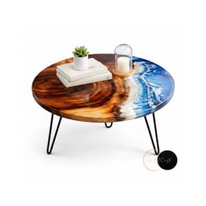 Puede incluir: Mesa de centro redonda con un diseño único. La mesa presenta un patrón de vetas de madera en una mitad y un diseño de resina azul y blanco que se asemeja a olas del océano en la otra. Tiene patas de horquilla negras y está decorada con una pequeña planta, libros y una vela.