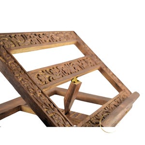 May include: A wooden book stand with intricate carvings. The stand has a hinged top that can be adjusted to different angles. The stand is made of dark brown wood and has a gold-colored metal hinge.