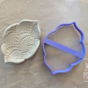 May include: A light-colored clay piece with a detailed lace-like pattern next to a purple plastic cookie cutter. The cutter has a decorative shape with a horizontal support. The text "WAIYI POTTERY" is visible in the lower right corner.