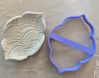 Può includere: Un pezzo di argilla colorata chiara con un motivo dettagliato a forma di pizzo accanto a un tagliabiscotti di plastica viola. Il tagliabiscotti ha una forma decorativa con un supporto orizzontale. Il testo "WAIYI POTTERY" è visibile in basso a destra.