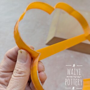 Pu&ograve; includere: Un utensile per ceramica arancione brillante, forse una stecca o uno strumento per modellare, &egrave; tenuto in mano. L'utensile ha una forma curva con un profilo piatto e largo. L'immagine include il testo "WAIYI POTTERY".