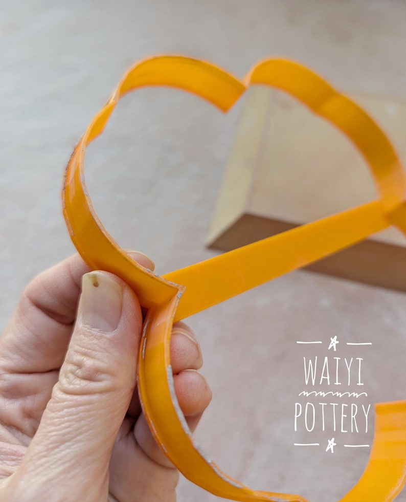 Pu&ograve; includere: Un utensile per ceramica arancione brillante, forse una stecca o uno strumento per modellare, &egrave; tenuto in mano. L'utensile ha una forma curva con un profilo piatto e largo. L'immagine include il testo "WAIYI POTTERY".