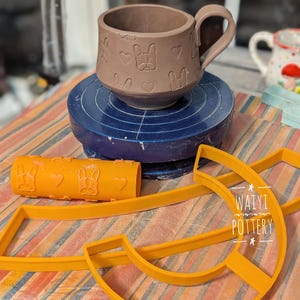 May include: A brown ceramic mug with a handle, decorated with a repeating pattern of hearts and dog faces. The mug is on a pottery wheel, and there are two orange plastic tools nearby. The tools are a rolling pin with a dog face design and a half-circle shape. The text "WAIYI POTTERY" is visible on one of the tools.