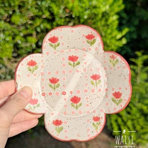 Pu&ograve; includere: Un sottobicchiere in ceramica bianca a forma di fiore con bordo rosso. Il sottobicchiere &egrave; decorato con fiori rossi e pois rosa. Il testo "WAIYI POTTERY" &egrave; visibile nell'immagine.