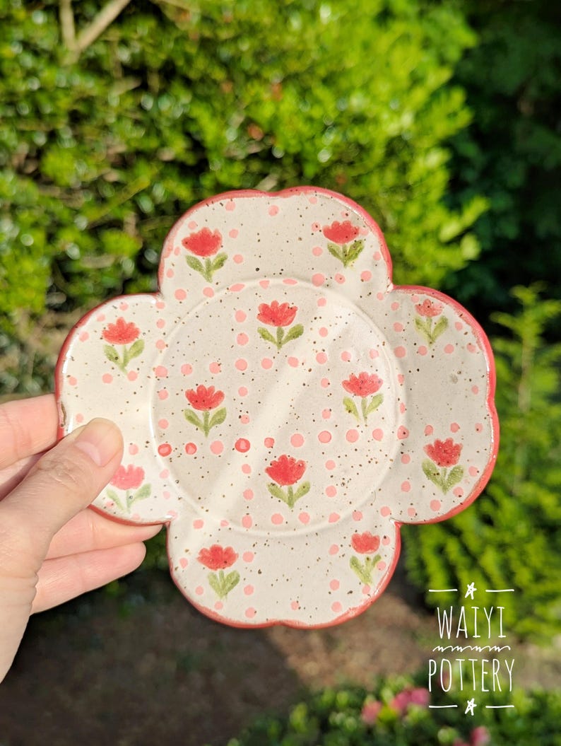 Pu&ograve; includere: Un sottobicchiere in ceramica bianca a forma di fiore con bordo rosso. Il sottobicchiere &egrave; decorato con fiori rossi e pois rosa. Il testo "WAIYI POTTERY" &egrave; visibile nell'immagine.