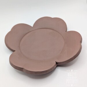 Pu&ograve; includere: Un piatto in ceramica a forma di fiore color malva. Il piatto ha un'area circolare centrale circondata da sei petali arrotondati. La superficie &egrave; liscia e opaca. Il piatto &egrave; probabilmente fatto a mano e adatto per servire cibo o come oggetto decorativo.