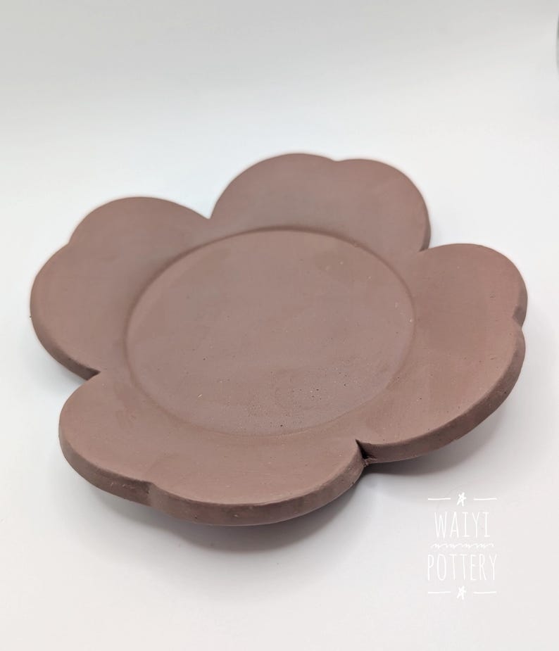 Pu&ograve; includere: Un piatto in ceramica a forma di fiore color malva. Il piatto ha un'area circolare centrale circondata da sei petali arrotondati. La superficie &egrave; liscia e opaca. Il piatto &egrave; probabilmente fatto a mano e adatto per servire cibo o come oggetto decorativo.