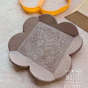 May include: A handmade, brown ceramic dish with a floral design in the center. The dish has a unique, scalloped edge. An orange, butterfly-shaped cookie cutter is in the background. The text "WAIYI POTTERY" is visible.