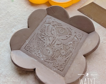 May include: A handmade, brown ceramic dish with a floral design in the center. The dish has a unique, scalloped edge. An orange, butterfly-shaped cookie cutter is in the background. The text "WAIYI POTTERY" is visible.