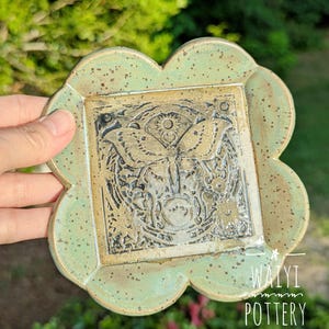 Pu&ograve; includere: Un piatto in ceramica fatto a mano con un centro quadrato e un bordo smerlato. Il centro presenta un disegno a farfalla dettagliato nei toni del marrone e del nero. Il piatto ha un bordo verde chiaro macchiettato e il testo "WAIYI POTTERY".