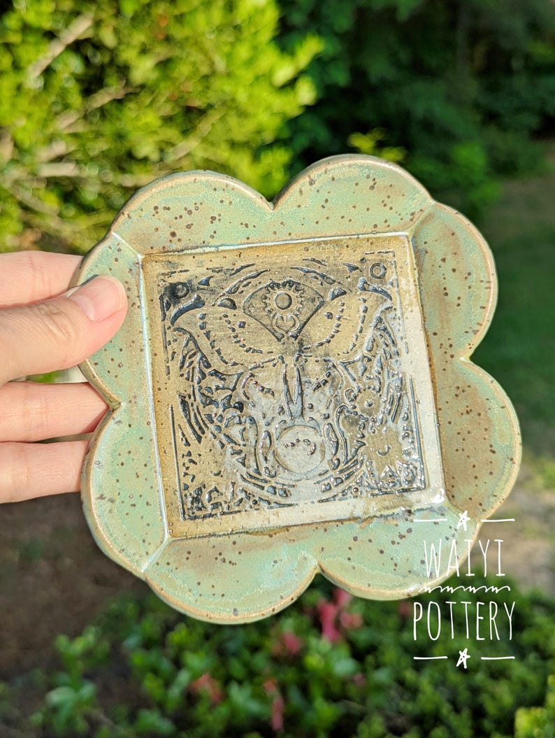 Pu&ograve; includere: Un piatto in ceramica fatto a mano con un centro quadrato e un bordo smerlato. Il centro presenta un disegno a farfalla dettagliato nei toni del marrone e del nero. Il piatto ha un bordo verde chiaro macchiettato e il testo "WAIYI POTTERY".