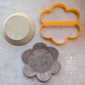 May include: A brown clay dish with a flower shape and a pattern of small dog faces and hearts. The dish is next to a yellow plastic flower-shaped cookie cutter.