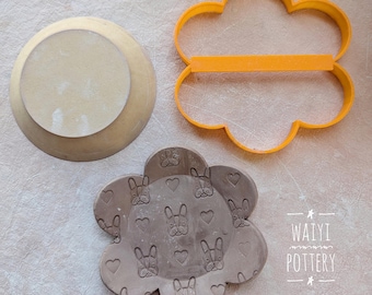 May include: A brown clay dish with a flower shape and a pattern of small dog faces and hearts. The dish is next to a yellow plastic flower-shaped cookie cutter.