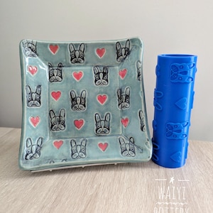 May include: A blue ceramic square dish with a pattern of red hearts and black and white French bulldog faces. A blue rolling pin with a similar pattern is next to the dish. The text "WAIYI POTTERY" is on the bottom of the image.
