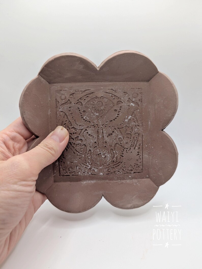 Pu&ograve; includere: Un sottobicchiere in ceramica marrone fatto a mano con un motivo floreale al centro. Il sottobicchiere ha una forma unica a quattro lobi. L'immagine include anche il testo "WAIYI POTTERY".