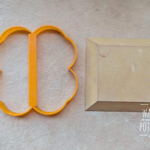 Pu&ograve; includere: Un tagliabiscotti arancione a forma di quadrifoglio e un blocco di legno quadrato sono esposti su una superficie di colore chiaro. Il tagliabiscotti ha una barra di supporto verticale. Il blocco di legno ha bordi smussati. Il testo "WAIYI POTTERY" &egrave; visibile.