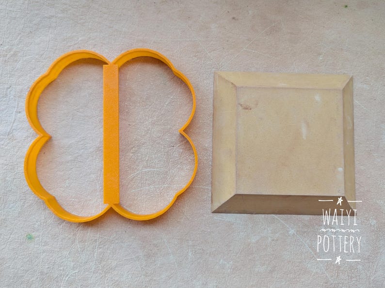 Pu&ograve; includere: Un tagliabiscotti arancione a forma di quadrifoglio e un blocco di legno quadrato sono esposti su una superficie di colore chiaro. Il tagliabiscotti ha una barra di supporto verticale. Il blocco di legno ha bordi smussati. Il testo "WAIYI POTTERY" &egrave; visibile.