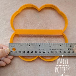 Pu&ograve; includere: Un tagliapasta in plastica arancione a forma di quattro lobi, con un righello che mostra una misura di circa 12,7 cm. Il tagliapasta &egrave; su una superficie di colore chiaro. Il testo "WAIYI POTTERY" &egrave; visibile.