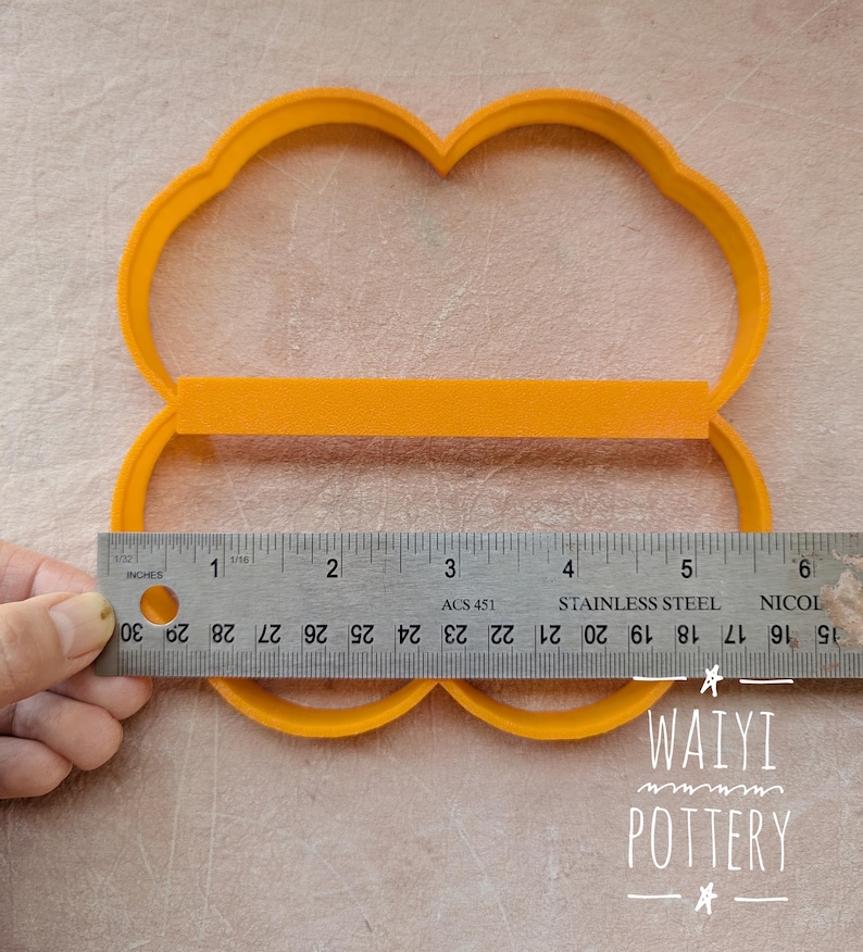 Pu&ograve; includere: Un tagliapasta in plastica arancione a forma di quattro lobi, con un righello che mostra una misura di circa 12,7 cm. Il tagliapasta &egrave; su una superficie di colore chiaro. Il testo "WAIYI POTTERY" &egrave; visibile.