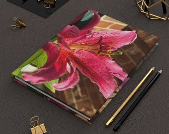 May include: A pink lily flower with green leaves on a brick wall background. The flower is in focus and the background is blurred. The image is printed on the cover of a notebook.