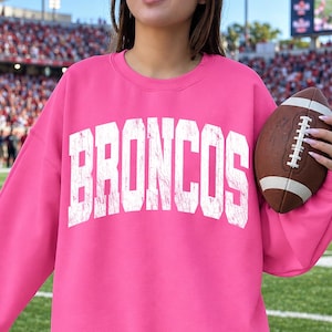 Puede incluir: Una sudadera de color rosa brillante de manga larga con la palabra BRONCOS impresa en letras mayúsculas blancas grandes y desgastadas en el pecho. La prenda tiene cuello redondo y se sostiene junto a un balón de fútbol americano.