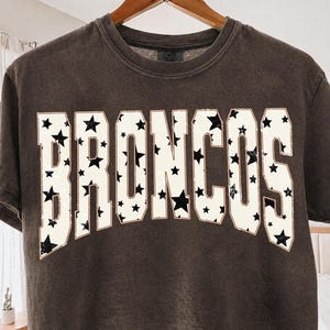 Puede incluir: Una camiseta de manga corta gris carbón con la palabra BRONCOS en letras grandes de bloque color blanco hueso. Las letras están rellenas con un patrón repetitivo de pequeñas estrellas negras y delineadas con un borde fino y claro.