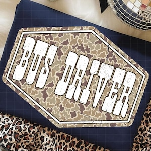 Könnte beinhalten: Marineblaues T-Shirt mit Tarnmuster und dem weißen Text "BUS DRIVER". Das Shirt ist auf einem Stoff mit Leopardenmuster gefaltet. Dekorative Discokugeln befinden sich im Hintergrund.