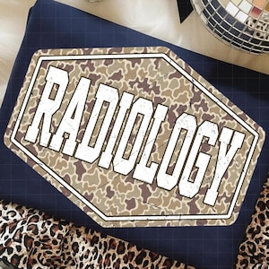 May include: A navy blue t-shirt with a camouflage-patterned patch that reads "RADIOLOGY" in bold white letters. The shirt is folded and placed on a white surface with disco balls and leopard print fabric.