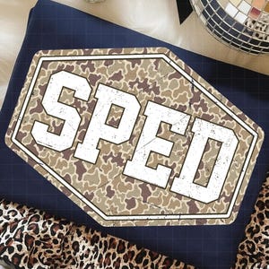 May include: Navy blue t-shirt with a distressed camouflage design and the word "SPED" in bold white letters. The shirt is folded and placed on a white surface with a leopard print fabric.