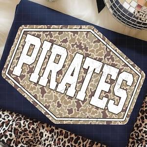 Puede incluir: Camiseta azul marino plegada con un gráfico desgastado de camuflaje que dice "PIRATES" en letras blancas. La camiseta está colocada sobre una tela con estampado de leopardo.