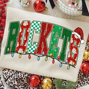 Könnte beinhalten: Cremefarbenes T-Shirt mit dem Wort "ROCKETS" in einem festlichen Design. Die Buchstaben sind grün, rot und weiß, mit Weihnachtsmann, Weihnachtsbäumen und Schachbrettmustern. Eine Lichterkette mit bunten Weihnachtslichtern verläuft darunter.
