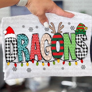 Op de afbeelding: Een lichtgrijs t-shirt met het woord "DRAGONS" in een feestelijk, kleurrijk ontwerp. De letters zijn versierd met patronen zoals buffelruit, luipaardprint en stippen, met vakantie-thema accenten zoals een kerstmuts en rendiergeweien. Een slinger van kerstverlichting loopt onder het woord.