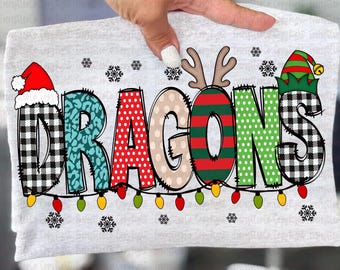 Puede incluir: Una camiseta gris claro con la palabra "DRAGONS" en un diseño festivo y colorido. Las letras están decoradas con patrones como cuadros de búfalo, estampado de leopardo y lunares, con acentos temáticos de vacaciones como un gorro de Papá Noel y cuernos de reno. Una cadena de luces navideñas corre debajo de la palabra.