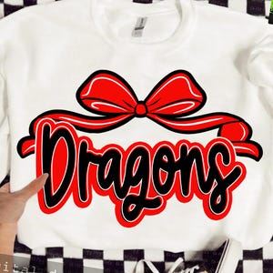 May include: White sweatshirt with a red bow and the word "Dragons" in a bold, red and black design. The text is in a playful, cursive font, making it a fun and eye-catching design. The sweatshirt is a casual, comfortable garment.