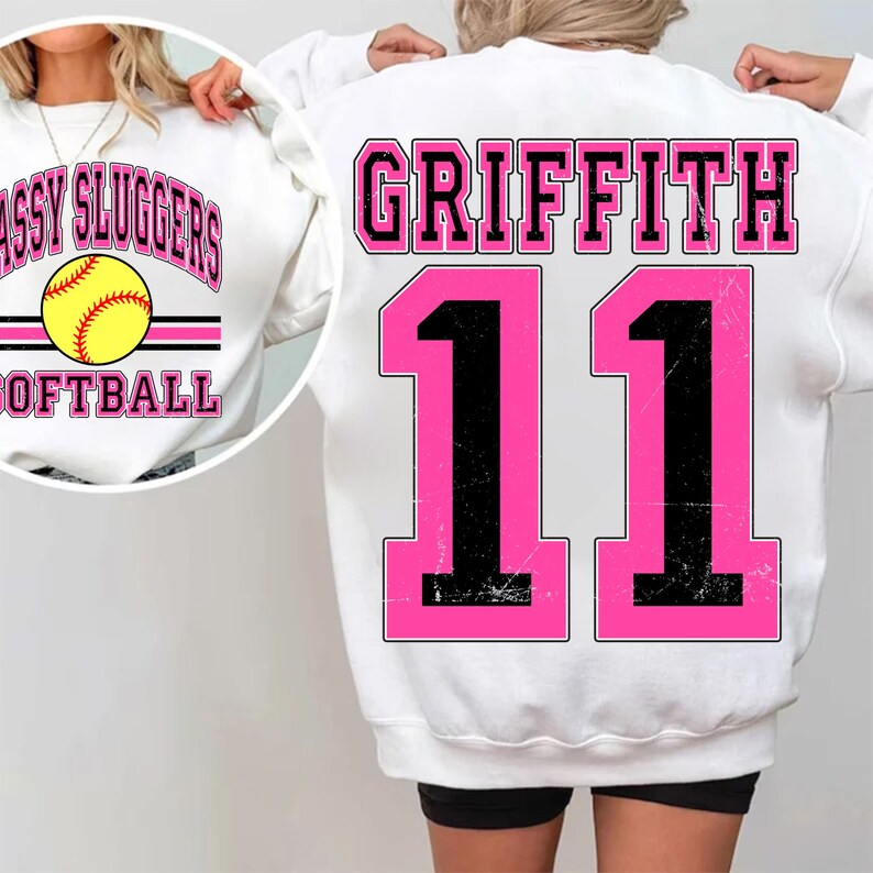 May include: White sweatshirt with pink and black lettering that says "Griffith 11". The front of the sweatshirt has a softball graphic with the text "Sassy Sluggers Softball".