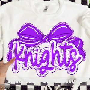 Pode incluir: Camisola branca com um grande laço roxo e a palavra "Knights" em uma fonte decorativa. A palavra "Knights" é preenchida com um padrão floral. O laço e as letras são delineados em branco.