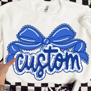 Puede incluir: Sudadera blanca con un gran lazo azul y la palabra "custom" en un diseño floral azul. El lazo tiene un ribete de encaje. La sudadera es de la marca Gildan. El diseño está centrado en la parte delantera.