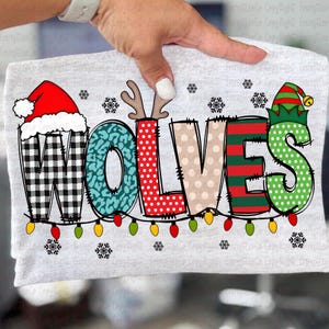 May include: A light gray t-shirt featuring the word "WOLVES" in a festive, holiday-themed design. The letters are decorated with various patterns, including buffalo plaid, teal leopard print, polka dots, and stripes. The design includes snowflakes and a string of colorful lights.