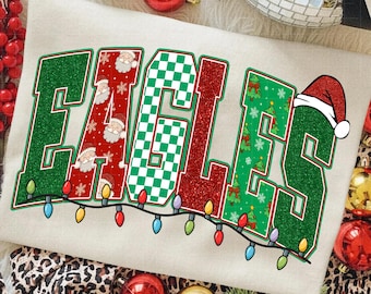 May include: Cream-colored sweatshirt with the word "EAGLES" in a festive design. The letters are outlined in green and filled with red glitter, green checkered patterns, and Christmas tree and Santa Claus motifs. A string of colorful lights and a Santa hat complete the holiday theme.