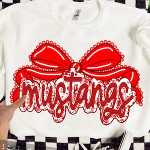 May include: White sweatshirt featuring a large red bow and the word "mustangs" in a decorative font. The text and bow have a floral pattern. The sweatshirt is a crew neck style.
