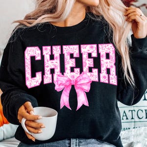 May include: Black sweatshirt with the word "CHEER" in large, pink, floral-patterned letters. A pink bow is centered below the word. The sweatshirt is a crew neck style.