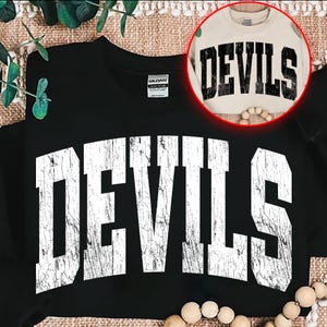 May include: Black sweatshirt with the word "DEVILS" in large, distressed white letters across the front. A smaller, tan sweatshirt with the same design is visible in a circular inset. The design has a vintage, collegiate aesthetic.