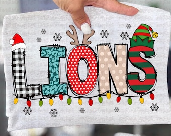 May include: A light gray t-shirt with the word "LIONS" in a festive design. The letters are decorated with buffalo plaid, teal leopard print, red polka dots, and tan polka dots. The "L" has a Santa hat, the "O" has antlers, and the "S" has an elf hat.