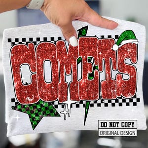 May include: A light gray t-shirt featuring the word "COMETS" in large, glittery red letters with a black outline. The design includes a green and black checkered star and a green Santa hat on the letter "S".