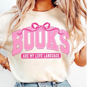 Pu&ograve; includere: Maglietta bianca con una grafica rosa che dice "BOOKS ARE MY LOVE LANGUAGE". La grafica presenta un fiocco rosa e la parola "BOOKS" in un font retr&ograve;.