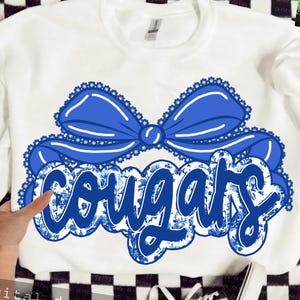 Peut inclure: Sweat-shirt blanc avec un grand nœud bleu et le mot "Cougars" dans un motif floral. Le texte est souligné en bleu et présente un motif floral. Le nœud a une bordure en dentelle.