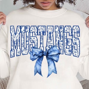 Puede incluir: Sudadera blanca con un dise&ntilde;o floral azul y blanco que deletrea "MUSTANGS" con un lazo azul en el centro.