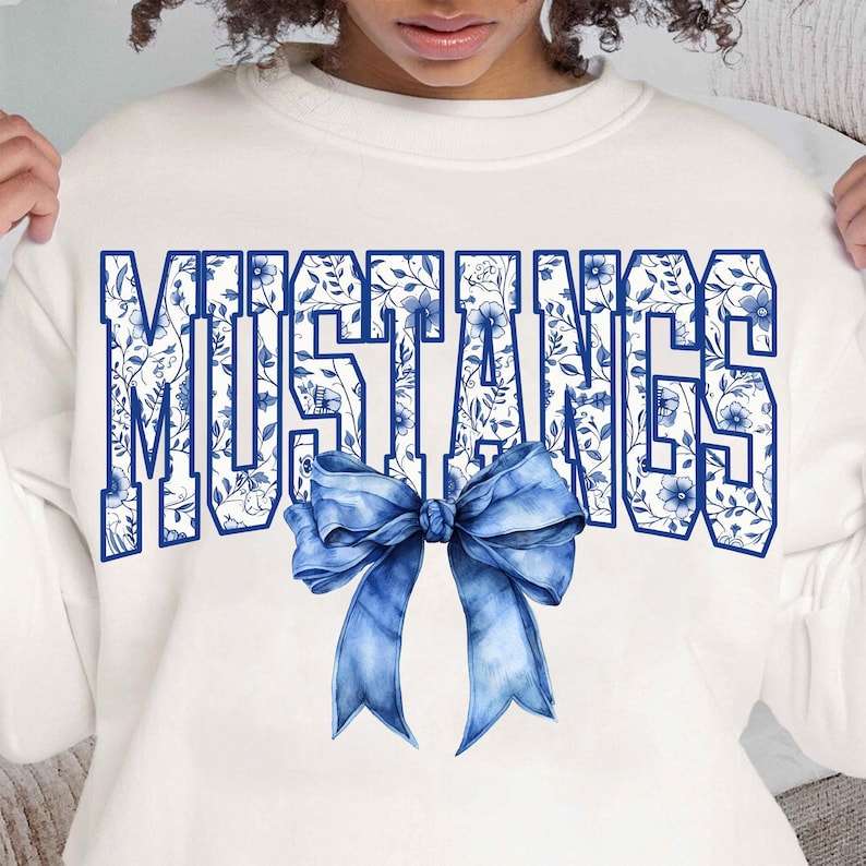 Puede incluir: Sudadera blanca con un dise&ntilde;o floral azul y blanco que deletrea "MUSTANGS" con un lazo azul en el centro.
