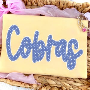 May include: A pale yellow t-shirt featuring the word Cobras in a blue font with a white polka dot pattern. The text is written in a cursive script style with a white outline. The shirt is folded and placed inside a woven basket with pink fabric.